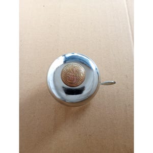 May include: A polished chrome bicycle bell with a small lever for ringing. The bell features a circular, gold-toned centre with the word "SIMPLEX" in a decorative font. The bell is round and has a classic design.