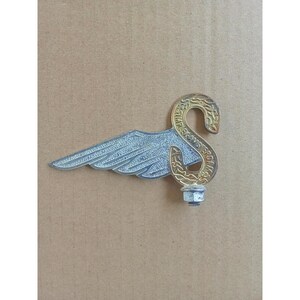 May include: A silver-toned metal wing emblem featuring a gold-toned curved neck section with the embossed text SIMPLEX AMSTERDAM. The piece includes a threaded bolt and nut at the base for attachment, displaying a textured finish on the wing surface.
