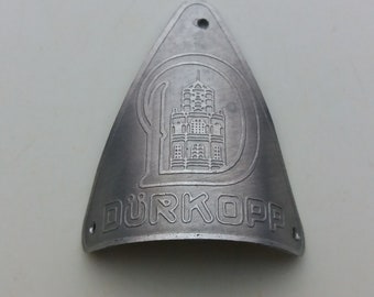 May include: A silver metal triangle-shaped badge with a stylized building and the text "Dürkopp" engraved on it.