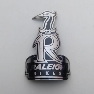 Op de afbeelding: Een zilver- en zwartkleurig metalen fietsbalhoofdplaatje met een gestileerd reiger-icoon aan de bovenkant, een grote letter R in het midden en de tekst RALEIGH BIKES in een vet, schreefloos lettertype op een gebogen banner onderaan.