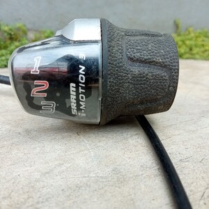 May include: A black and silver SRAM 1-Motion rear derailleur shifter with a rubber grip. The shifter has numbers 1, 2, and 3 printed on it.