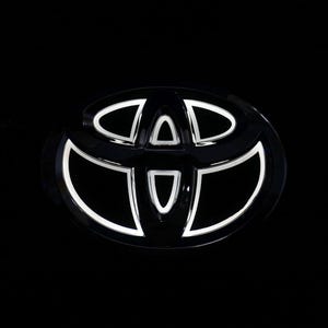 May include: A black Toyota emblem featuring a glossy finish with integrated white LED lighting that outlines the three overlapping elliptical shapes of the logo, set against a solid black background to highlight the illuminated contours.