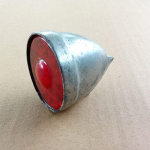 May include: A vintage bicycle tail light featuring a red lens and a silver metal casing. The lens is circular with a central, raised red detail. The casing has a conical shape with a small, pointed end.