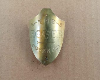 Könnte beinhalten: Ein schildförmiges Fahrrad-Steuerkopfschild aus Messing mit der eingravierten Aufschrift THE ROVER COVENTRY. Die Metallplatte hat eine spitz zulaufende Unterseite, eine geschwungene Oberseite und zwei kleine, vertikal zentrierte Montagelöcher.