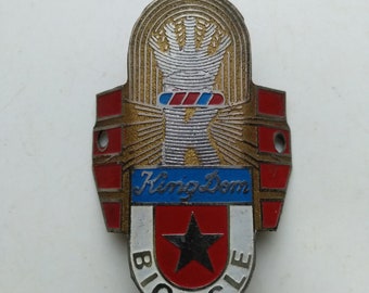 May include: A vintage bicycle head badge with a gold, red, and white design. The badge features a stylized hand holding a barber pole and the text "Kingdom Bicycle".