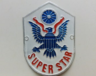 May include: A white metal shield with a blue eagle and red ribbon, a blue wheel, and the text "SUPER STAR" in red letters.