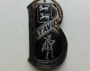 Könnte beinhalten: Ein Vintage-Metallabzeichen in Form des Buchstabens "B". Das Abzeichen hat einen schwarzen Hintergrund mit dem Wort "BATAVUS" und einem Wappen-Design. Das Abzeichen ist silberfarben.