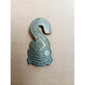 Può includere: Un emblema vintage in metallo a forma di S stilizzata, con una figura con un'arpa e le parole "SUPER CYCLE". L'emblema ha una finitura bronzata ed è probabilmente un distintivo o un logo.