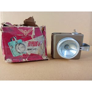 May include: A vintage brown metal bicycle headlight with a silver reflector and a small top switch, positioned next to its original worn red cardboard box featuring the text Flying Pigeon Bicycle and a graphic illustration of the light.