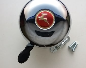 May include: A chrome bicycle bell with a red and gold deer logo. The bell is attached to a silver bracket with two screws.