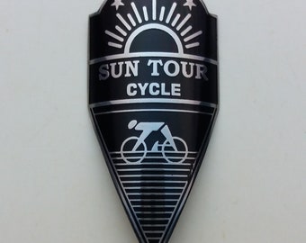 Könnte beinhalten: Schwarzes und silbernes Fahrrad-Kopfzeichen mit einer stilisierten Sonne und fünf Sternen, dem Text "SUN TOUR CYCLE" und einer Silhouette einer Person, die Fahrrad fährt.