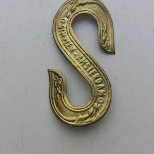 May include: A gold-coloured metal hook with the inscription "Simplex Amsterdam" around the outside edge.