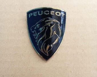 Könnte beinhalten: Ein schwarzes und goldenes Peugeot-Emblem mit dem Markennamen und einem Löwenlogo. Das Emblem ist schildförmig und hat eine glänzende Oberfläche. Der Löwe ist im Profil dargestellt, mit offenem Maul.