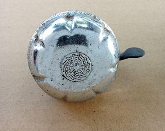 May include: A vintage silver bicycle bell with a black lever. The bell has a circular design with a decorative pattern in the center. The bell is made of metal and shows signs of wear.
