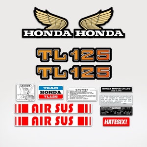 Puede incluir: Un conjunto de calcomanías de motocicleta vintage en negro, naranja, blanco y rojo. Las calcomanías presentan las palabras "Honda", "TL125", "Air Sus" y "Team Honda TL125". También hay una etiqueta de precaución con instrucciones para instalar un elemento de filtro de aire.