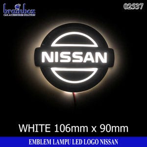 May include: A circular Nissan car emblem featuring a white LED backlight. The logo displays the word NISSAN in white lettering across a horizontal bar within a circular frame. The emblem measures 106mm by 90mm and includes a red wire connection.
