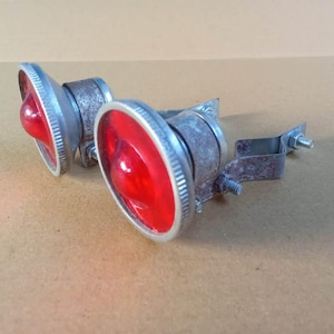 May include: Two circular red glass reflectors with silver-toned metal rims and textured housings. Each reflector features an integrated metal mounting bracket with a threaded bolt and nut, designed for attachment to a frame or surface.