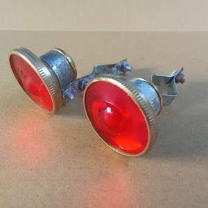 May include: Two circular red reflectors with gold-toned metal rims and silver-toned metal mounting brackets. Each reflector features a textured edge and a central dome, connected by a central metal bar with visible screws and hardware components.