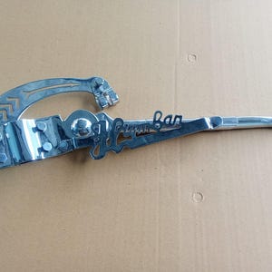 May include: A chrome guitar tremolo arm, featuring the word "Fender" in a stylized script. The arm has a curved handle and a mounting plate with screws. The design includes arrow-like cutouts. The tremolo arm is made of chrome.