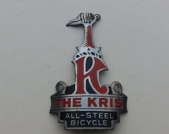 Könnte beinhalten: Ein silbernes Fahrrad-Kopfzeichen mit einem stilisierten roten und schwarzen Buchstaben "K" und dem Text "The Kris All-Steel Bicycle".