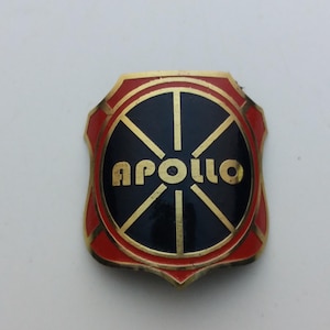 Puede incluir: Una insignia de metal dorada y roja con un diseño de rueda negro y dorado y la palabra "APOLLO" en letras doradas.