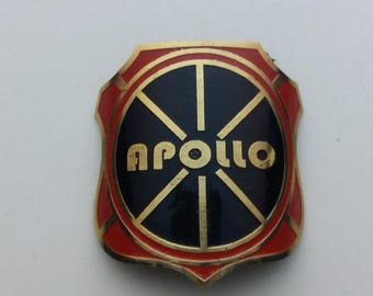 May include: A gold and red metal badge with a black and gold wheel design and the word "APOLLO" in gold lettering.