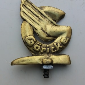 May include: A vintage brass emblem featuring a stylized hand with a wing design. The word "Göricke" is inscribed within the emblem. The emblem is mounted on a base with a threaded stud and nut.