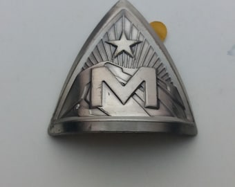 May include: A silver metal badge with a mountain range and a star above the letter "M".