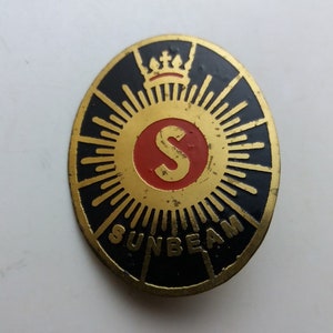 May include: Vintage Sunbeam badge. The oval badge features a gold sunburst design with a black border. The center has a red circle with the letter "S" and the word "SUNBEAM" at the bottom. A crown is at the top.