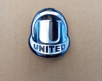 May include: A vintage metal emblem with a black and silver design. The emblem features a large silver "U" inside a black shield shape, with the word "UNITED" in white lettering at the bottom. The emblem is likely from a vehicle or machine.
