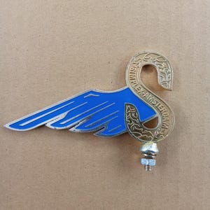 May include: A vintage metal emblem with a blue wing design and silver trim, and a gold-coloured, stylised letter "S" shape. The text "SIMPLEX-AMSTERDAM" is inscribed on the gold element. A bolt and nut are attached to the bottom.