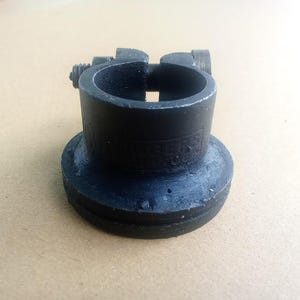 May include: A black metal clamp with a cylindrical body and a flared base. The clamp features an adjustment screw and a slot for securing an object. The base has a circular design. The clamp is likely used for holding or connecting items.