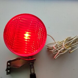 Op de afbeelding: Een cirkelvormig rood licht met een getextureerd, gefacetteerd lensoppervlak. Het licht is gemonteerd op een zilverkleurige metalen beugel en is verbonden met een opgerolde witte elektrische draad die eindigt in een rode krokodillenklem.