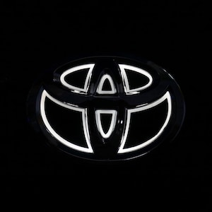 Op de afbeelding: Een Toyota-voertuigembleem met een glanzend zwart ovaal frame met drie overlappende elliptische vormen aan de binnenkant. De binnenranden van het logo zijn verlicht met een helder wit licht, wat een scherp contrast vormt met de donkere achtergrond.