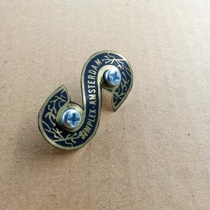 May include: A brass-toned metal pin in a stylized S-shape, featuring black enamel detailing and the text "SIMPLEX-AMSTERDAM". The pin is secured with two silver screws. Decorative flourishes are visible on the edges.