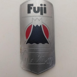 Puede incluir: Una placa de metal plateado para el tubo de dirección de una bicicleta con la palabra Fuji en texto negro en la parte superior. Debajo del texto hay un gráfico de una montaña en blanco y negro sobre un fondo circular rojo. La palabra Cycling está grabada.