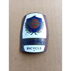Könnte beinhalten: Vintage Right Way Fahrrad-Emblem. Das schildförmige Emblem hat einen weißen Hintergrund, rote Verzierungen und ein blau-schwarzes zentrales Design. Die Texte "Right Way" und "BICYCLE" sind sichtbar.