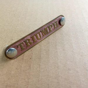 Könnte beinhalten: Ein rechteckiges Triumph-Emblem mit dem Wort "TRIUMPH" in goldfarbenen Buchstaben. Das Emblem hat einen kupferfarbenen Hintergrund und ist mit zwei silbernen Nieten befestigt. Wahrscheinlich für ein Motorrad oder ein klassisches Auto.