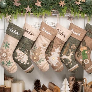 May include: Six personalized Christmas stockings in shades of beige, brown, and green hang from a white mantel. Each stocking is embroidered with a name and a festive design, including snowflakes, a bear, a snowman, a Christmas tree, a deer, and flowers.