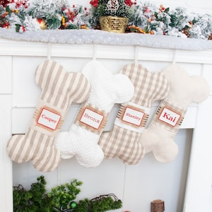 May include: Four personalized dog bone-shaped Christmas stockings hanging on a white fireplace mantel. The stockings are made of beige fabric with different patterns: stripes, gingham, and a white textured fabric. The stockings are labeled with the names "Cooper", "Bronx", "Stanley", and "Kai" in red lettering.