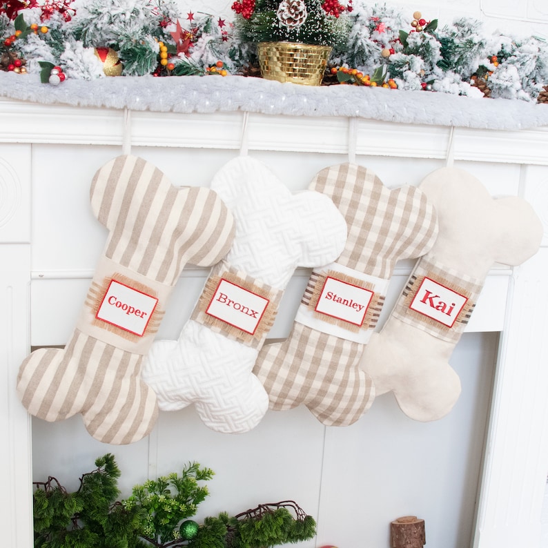 May include: Four personalized dog bone-shaped Christmas stockings hanging on a white fireplace mantel. The stockings are made of beige fabric with different patterns: stripes, gingham, and a white textured fabric. The stockings are labeled with the names "Cooper", "Bronx", "Stanley", and "Kai" in red lettering.
