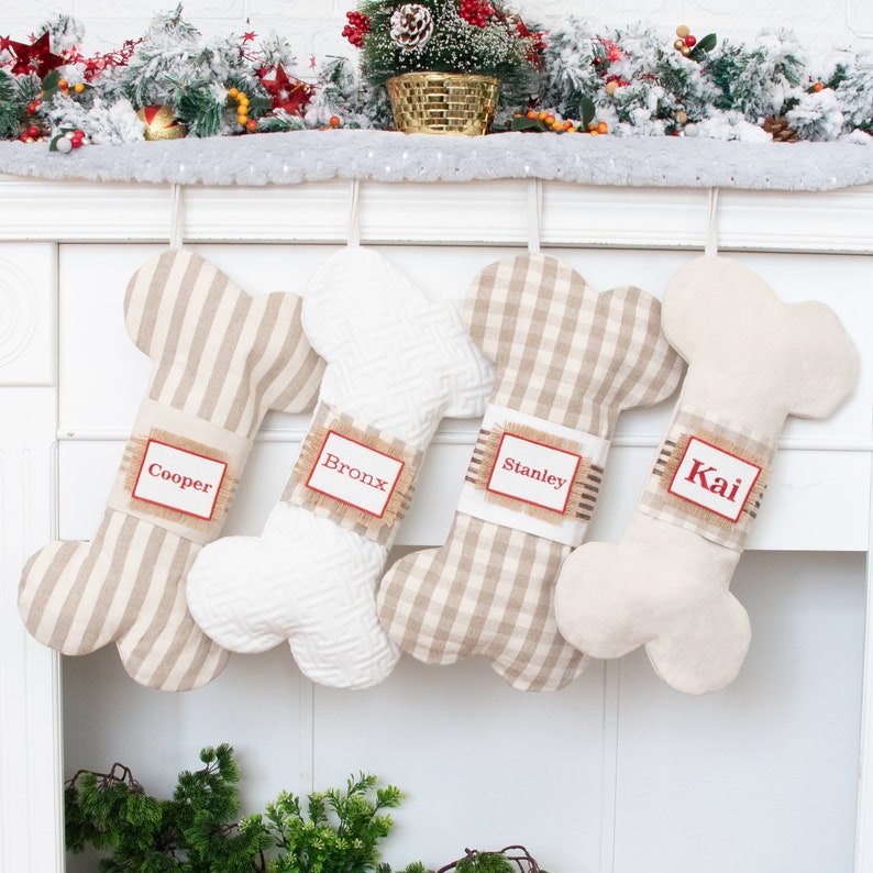 May include: Four beige and white dog bone-shaped Christmas stockings with names "Cooper", "Bronx", "Stanley", and "Kai" embroidered on them.