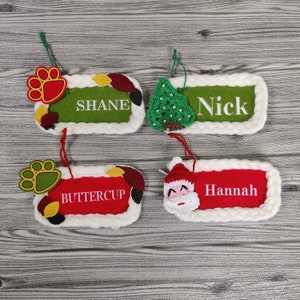 May include: Four personalized Christmas ornaments with white trim. Each ornament features a different design: a paw print, a Christmas tree, Santa Claus, and autumn leaves. The ornaments are labeled with the names "Shane", "Nick", "Buttercup", and "Hannah".