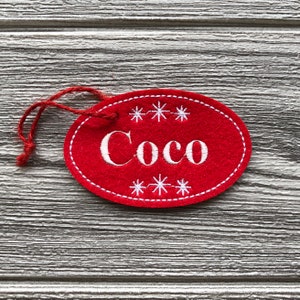 May include: Red felt oval ornament with white embroidery. The ornament has the name "Coco" and snowflakes around the name.