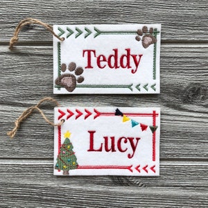 May include: Two rectangular white felt ornaments with embroidered names "Teddy" and "Lucy". "Teddy" has paw prints and green arrow border. "Lucy" has a Christmas tree and a red arrow border. Both have twine loops.