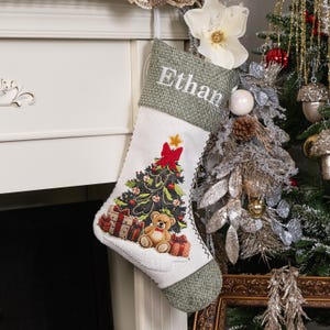 Puede incluir: Un calcet&iacute;n navide&ntilde;o con el nombre "Ethan" bordado en blanco sobre una parte superior de arpillera verde. El calcet&iacute;n presenta un dise&ntilde;o de &aacute;rbol de Navidad con un lazo rojo, adornos y un oso de peluche. Los regalos est&aacute;n en la base del &aacute;rbol.