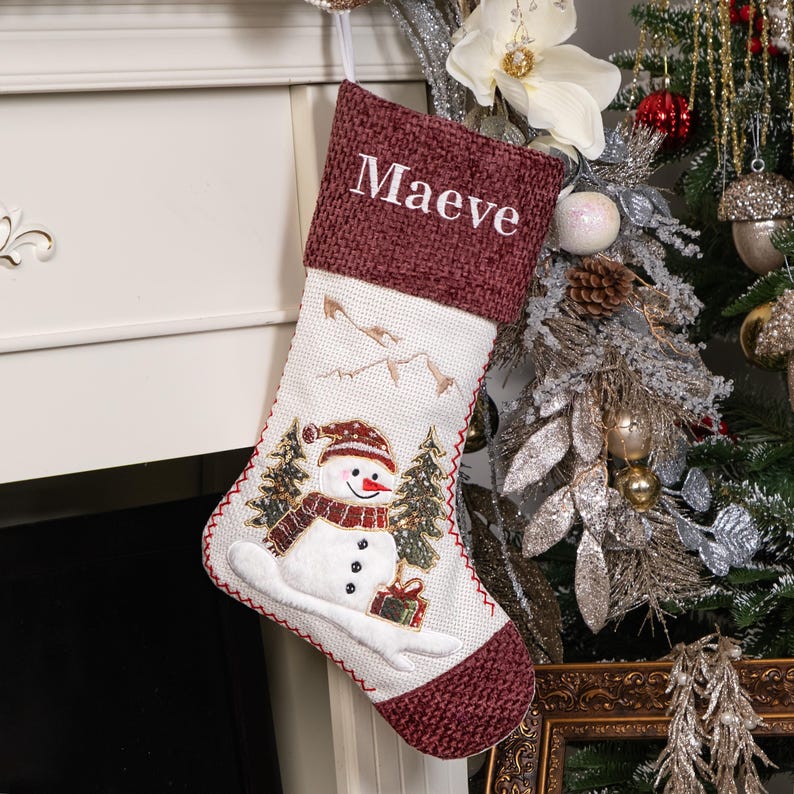 Puede incluir: Un calcet&iacute;n navide&ntilde;o con pu&ntilde;o burdeos y el nombre "Maeve" bordado en blanco. El cuerpo del calcet&iacute;n presenta un dise&ntilde;o de mu&ntilde;eco de nieve con &aacute;rboles y monta&ntilde;as. Costuras rojas delinean los bordes.