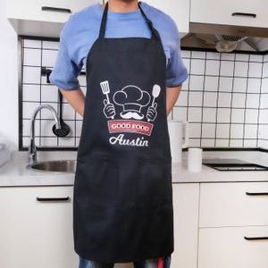 May include: Black apron with a white chef hat, mustache, and crossed utensils. The text "Good Food Austin" is printed on the apron.