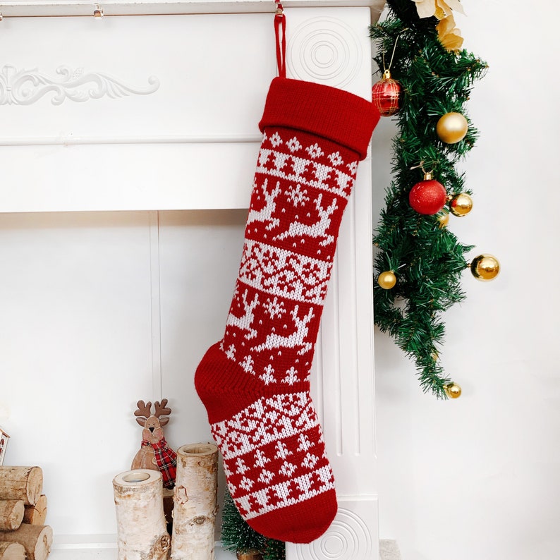 May include: A red Christmas stocking with a white reindeer and snowflake pattern. The stocking is hanging from a white mantelpiece, with a green garland and ornaments in the background. The stocking is made of knitted fabric.