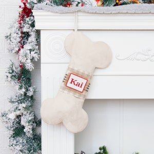 May include: A beige bone-shaped Christmas stocking with a red and white plaid ribbon and a personalized name tag that says "Kai".